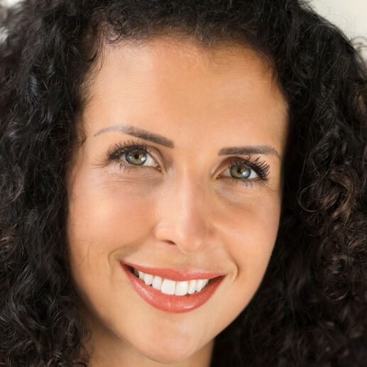Woman with curly dark hair, hazel eyes, and pink lipstick, smiling and looking at the camera.