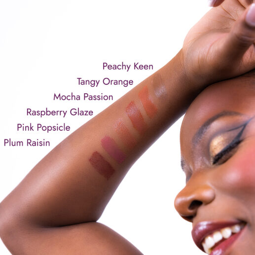 Lip color swatches on a woman's arm labeled: Peachy Keen, Tangy Orange, Mocha Passion, Raspberry Glaze, Pink Popsicle, and Plum Raisin. The woman is smiling with her eyes closed.